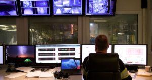 Person i arbeidsklær sitter i et kontrollrom i en fabrikk, foran kontrollskjermer og overvåkingsskjermer.