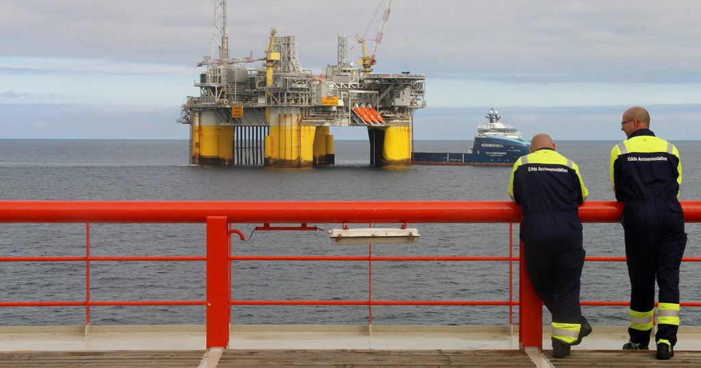 To arbeidstakarar i arbeidsklede står på eit skipsdekk og ser mot ein oljeinstallasjon i Nordsjøen.