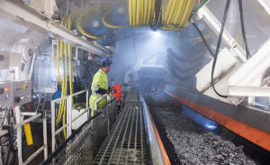 Operatør i arbeidsklede står ved eit transportbelte på tunnelboremaskina, som fraktar knust stein frå borehovudet.