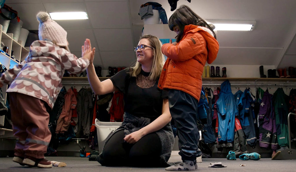En barnehageansatt gir en "high five" til to barn med ytterklær på.