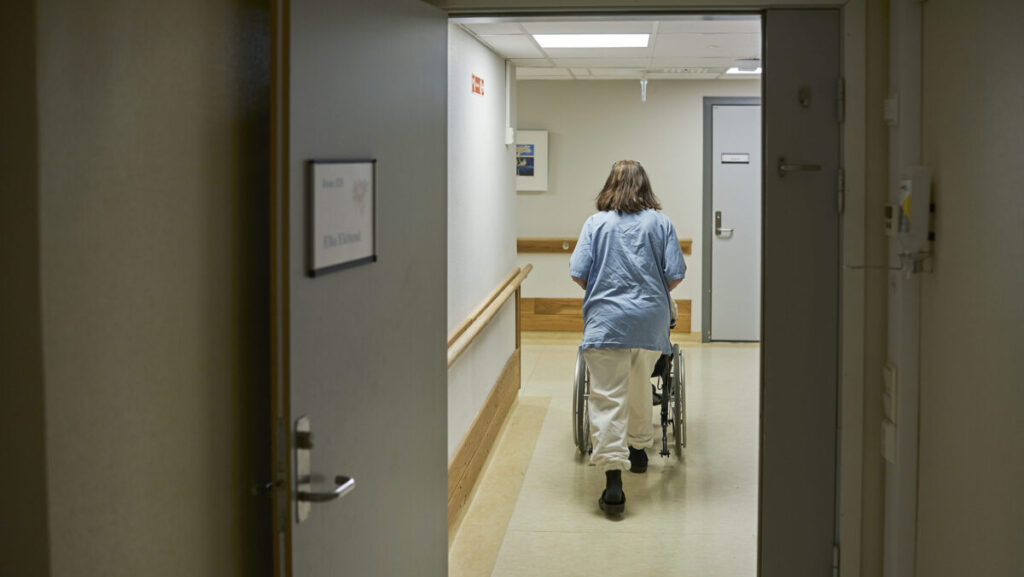 Sjukepleiar trillar ein rullestol i en sjukeheimsgang.