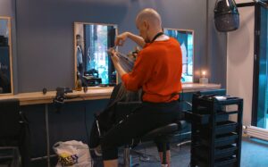 Ein mannleg frisør sit på ein høg krakk og arbeider med håret til ein kunde, som sit i frisørstolen.