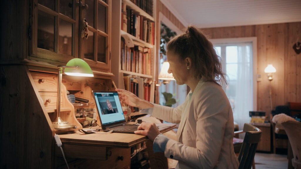Kvinne sit ved ein pult i ei stove, og deltar på zoom-møte på ein bærbar PC.