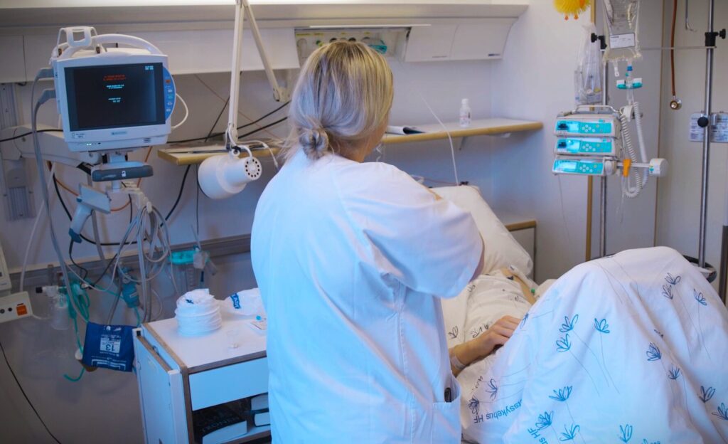 Sjukepleiar arbeider ved sida av ein pasient i sjukehusseng.