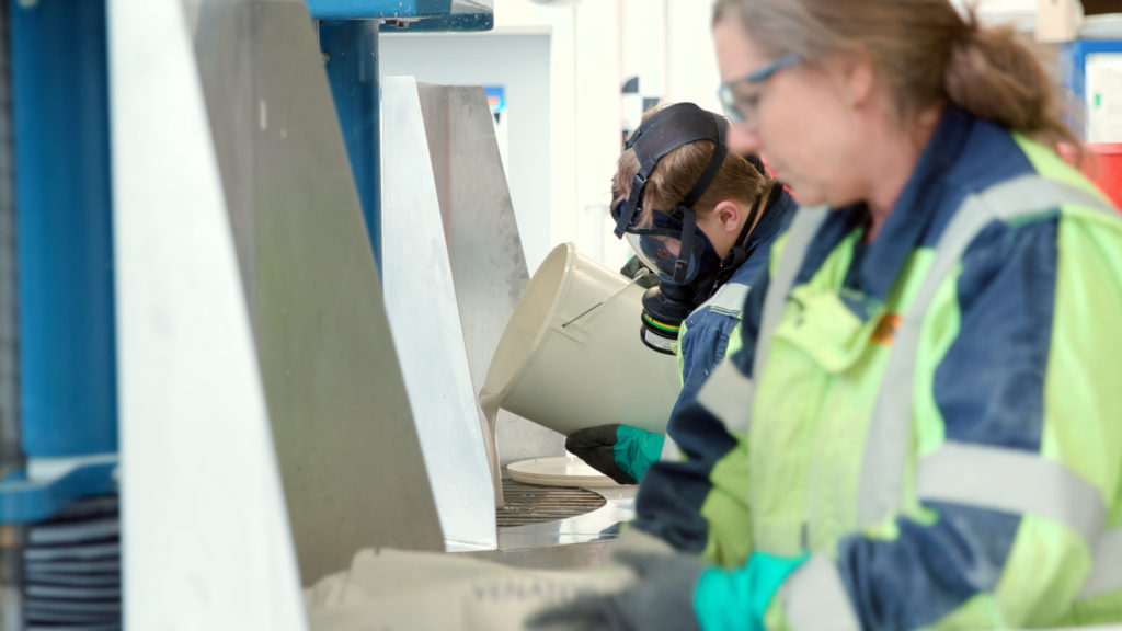 En kvinne og en mann i arbeidsklær i et industrilokale, heller flytende væske i en maskin fra en bøtte.