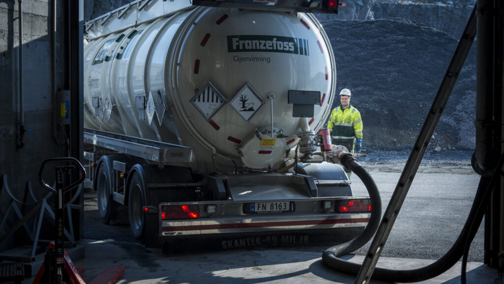 Ein sjåfør står ved sida av ein lastebil med slamtank, som tømmer slam gjennom ein stor slange.