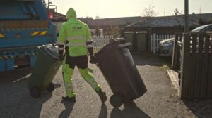 Person i arbeidsklede trillar to avfallscontainerar mot ein avfallsbil.