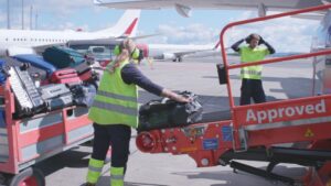 To personar i arbeidsklede og refleksvest flyttar bagasje frå vogner til fly, på ein flyplass.