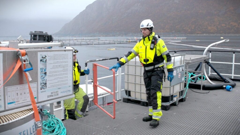 To operatørar på ei oppdrettsmerde, på sjøen ved kysten, i grått haustvêr.