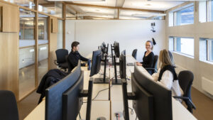 Tre kollegaer sitter ved sine PC-skjermer og snakker sammen, i et felles kontor med flere tomme kontorplasser.