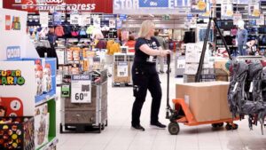 Kvinneleg butikktilsett i eit stort butikklokale flyttar ei pappkasse med ei tralle.