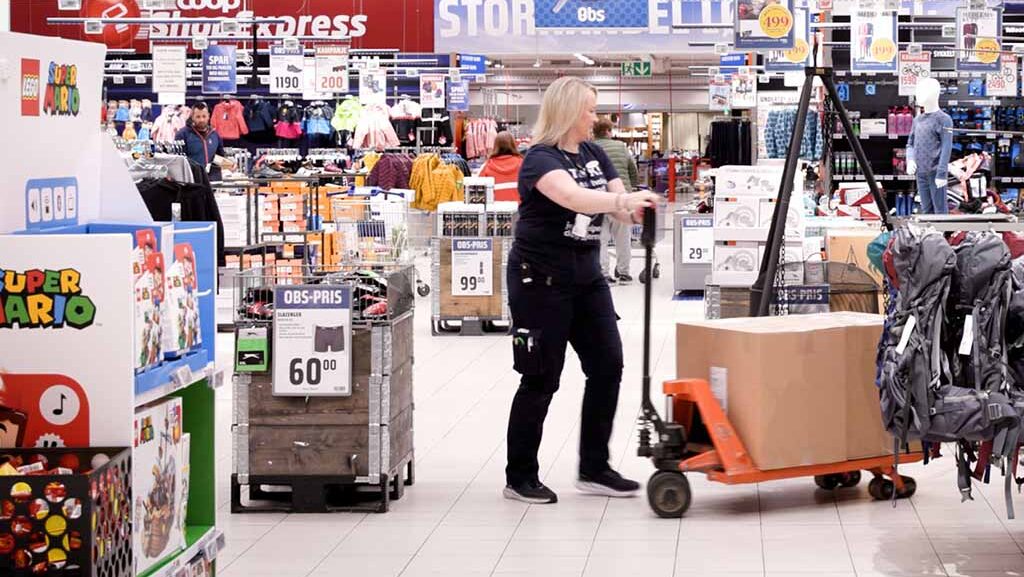 Kvinneleg butikktilsett i eit stort butikklokale flyttar ei pappkasse med ei tralle.