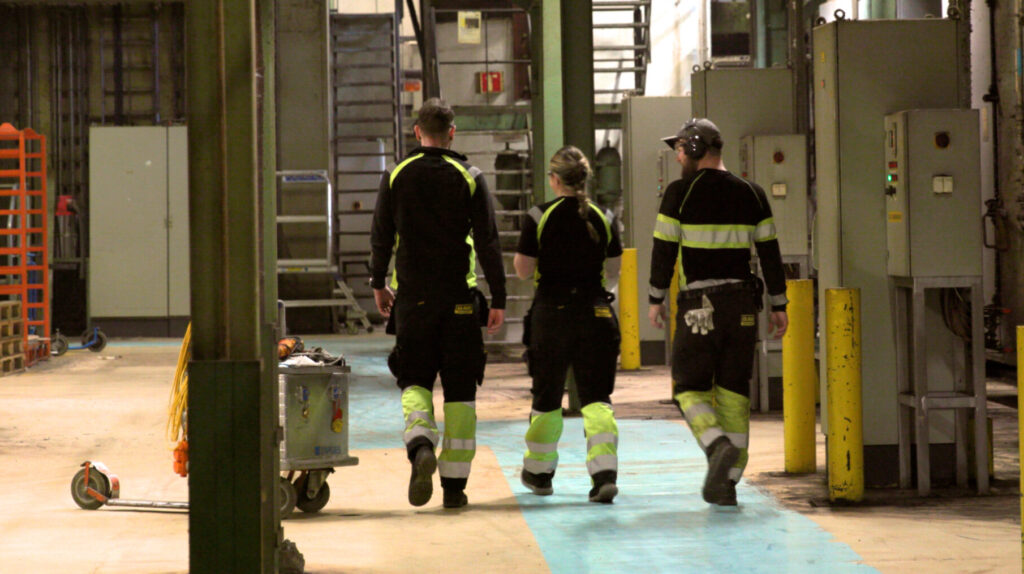 Tre kollegaer går i et industrilokalet og snakker hyggelig sammen.