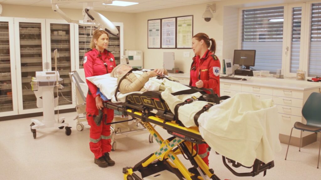 To ambulansearbeidere står ved en pasient på båre, på et sykehus