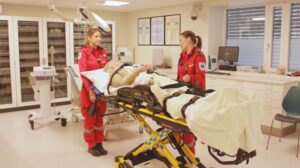 To ambulansearbeidere står ved en pasient på båre, på et sykehus