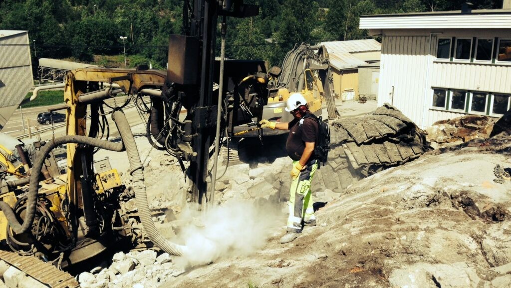 Mann med arbeidsklær og hjelm står ved en bergboremaskin, med synlig steinstøv fra borret